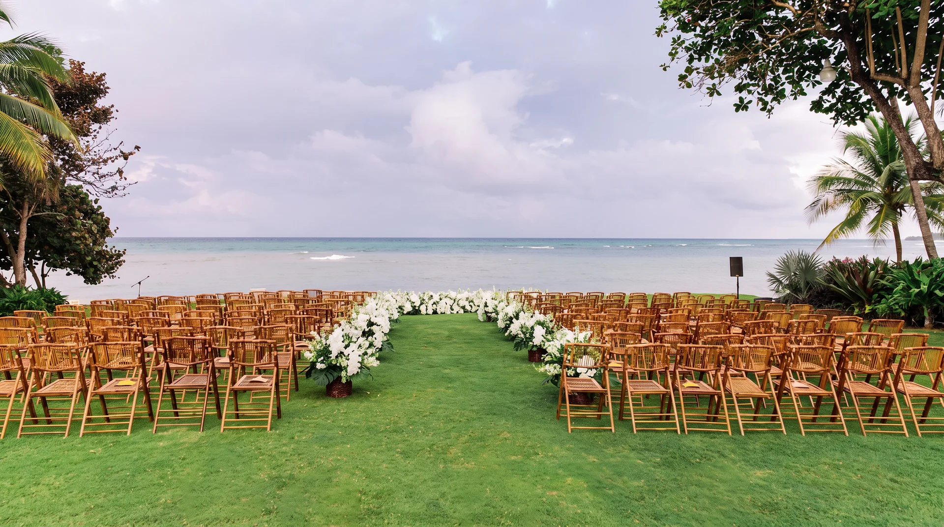 A ceremony lawn at the water's edge — bamboo chairs and a white-flower aisle facing the sea